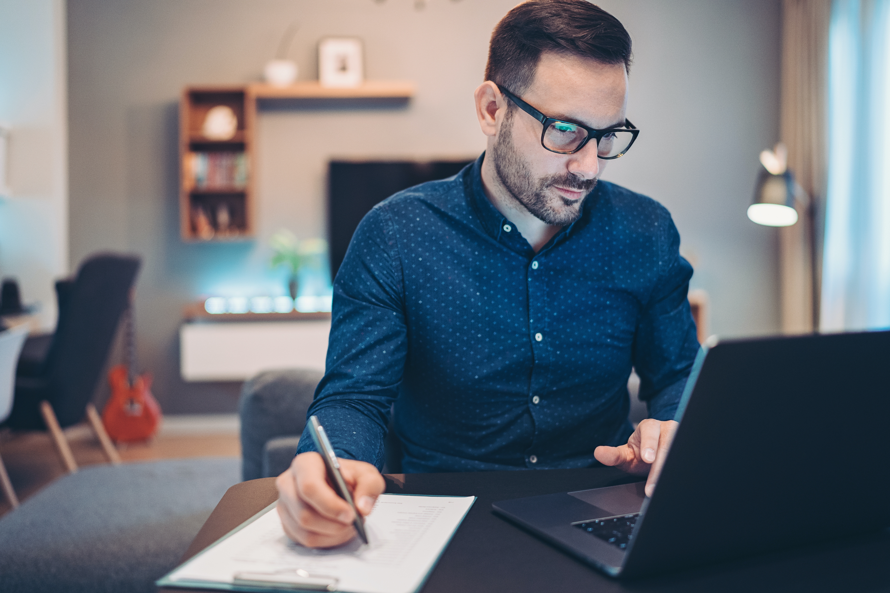 A man takes notes on paper while looking at his laptop